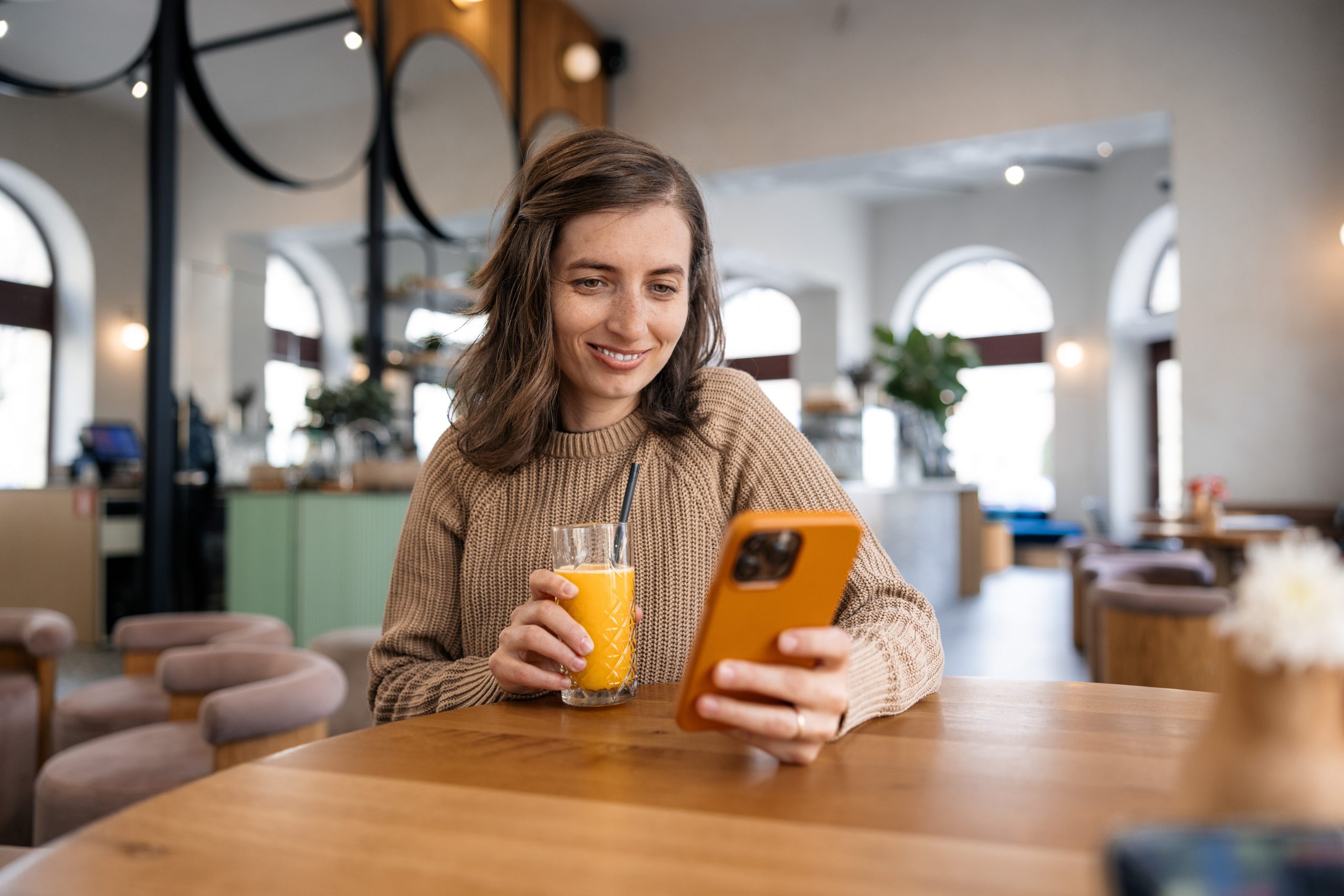 Woman smiling at her phone in a café while holding a glass of orange juice, as if reading or commenting on social media.