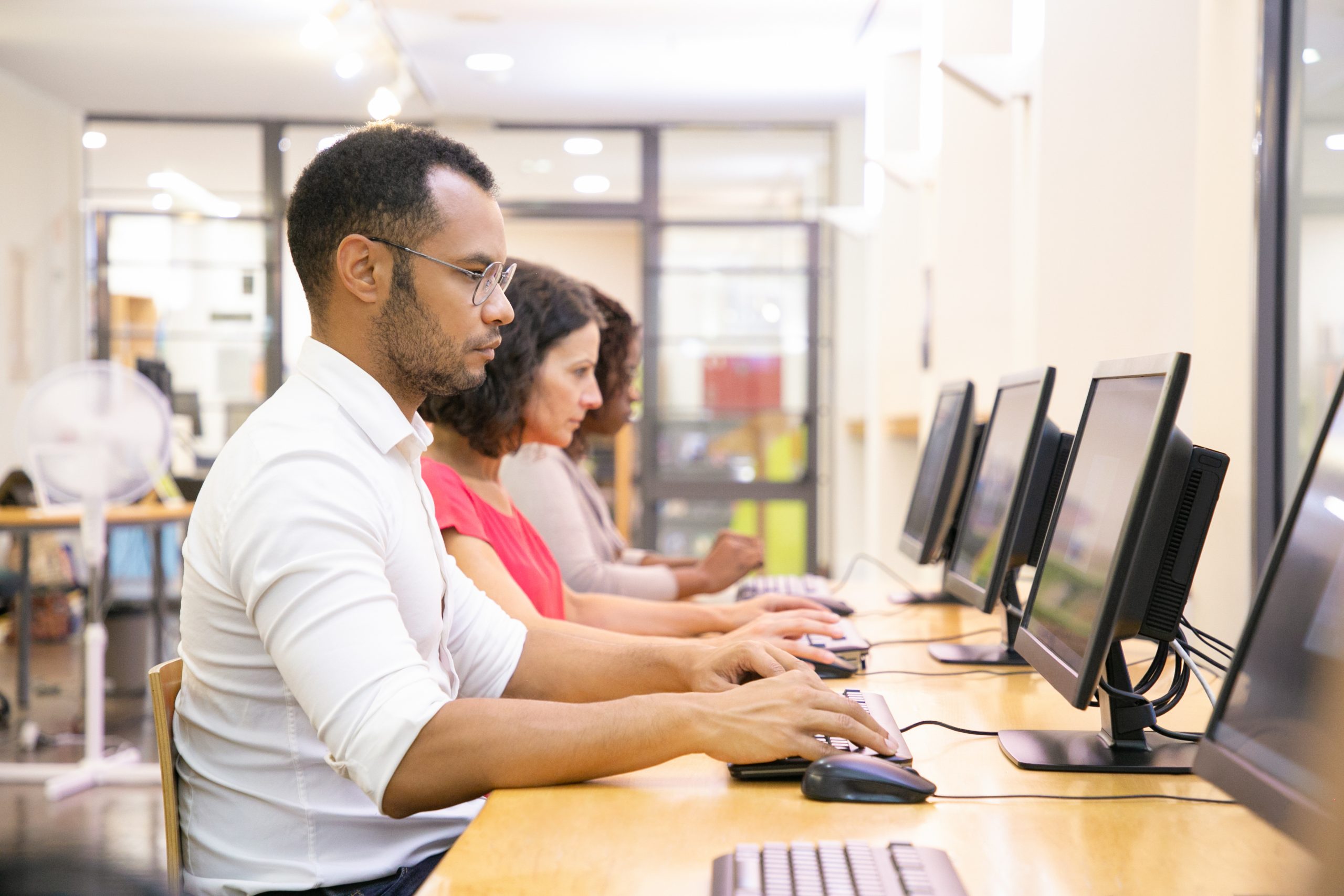 Three adults sit in a row at desktop computers in a bright, modern computer lab, with a man in the foreground focused on typing.